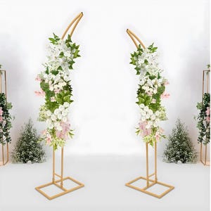 以下が含まれることがあります： 白、ピンク、緑の花の装飾が施された2つの金色の金属製アーチスタンド。スタンドは正方形のベースと湾曲した上部を備え、エレガントなディスプレイを作成します。結婚式やイベントに最適です。