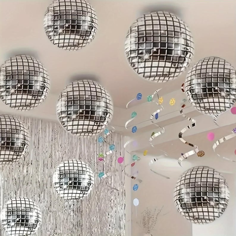 May include: Several silver disco balls are suspended from a white ceiling, reflecting light. The spheres are covered in small, mirrored squares. Decorative streamers and confetti add to the festive atmosphere, suggesting a party or celebration.