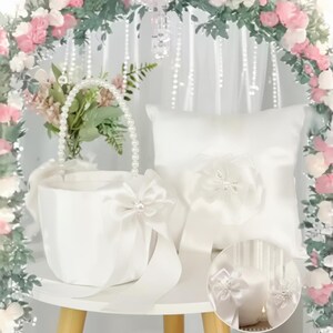 Puede incluir: Una cesta de boda de satén blanco con asa y lazo de perlas, y una almohada para anillos a juego con un adorno floral. Los artículos se exhiben sobre una pequeña mesa blanca, con un arco floral en el fondo.
