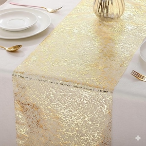 Puede incluir: Camino de mesa dorado con un delicado diseño similar a una telaraña. El camino de mesa está colocado sobre un mantel blanco, con cubiertos dorados y platos blancos en el fondo. Añade un toque de elegancia a la mesa.