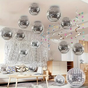 May include: A room decorated with multiple silver disco balls of varying sizes, hanging from the ceiling. Silver fringe and colorful streamers add to the festive atmosphere. A sofa and wooden furniture are visible in the background.