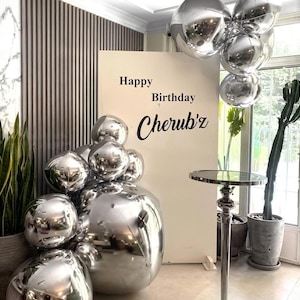 Puede incluir: Una escena de fiesta de cumpleaños con globos plateados y un letrero que dice "Happy Birthday Cherub'z". Una pequeña mesa redonda está frente a una ventana. También se ven plantas en macetas y un cactus.