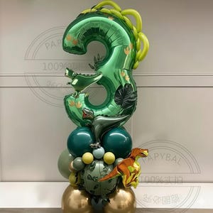 May include: A green number 3 foil balloon with a dinosaur design, part of a balloon arrangement. The arrangement includes green, gold, and yellow balloons, and a small orange and yellow dinosaur balloon. Ideal for a third birthday celebration.