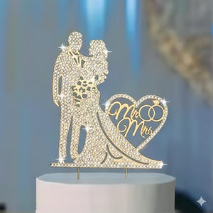 May include: A gold wedding cake topper depicting a couple embracing, embellished with rhinestones. The design includes a heart with the words "Mr & Mrs" and two rings. The topper is placed on a white cake.