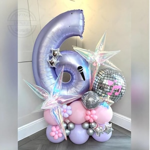Puede incluir: Un arreglo festivo de globos con un gran globo número 6 lavanda, rodeado de globos más pequeños en rosa, plateado y lavanda. Globos iridiscentes en forma de estrella y una bola de discoteca añaden brillo. Incluye un gráfico de micrófono.