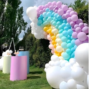 May include: A vibrant rainbow balloon arch with white, pink, yellow, blue, and purple balloons. The arch is set up outdoors with three colorful cylindrical pedestals in the foreground, perfect for a party or event.