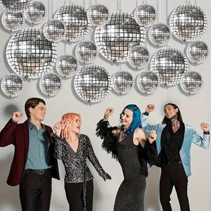 May include: A group of people dancing in front of a wall decorated with silver disco balls. The disco balls are round and reflective, with a grid-like pattern. The people are wearing various outfits, including a maroon blazer, a black dress, and a light blue jacket.
