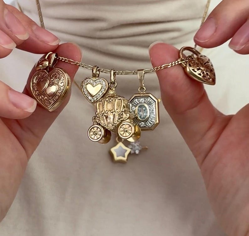 K&ouml;nnte beinhalten: Eine goldene Halskette mit verschiedenen Anh&auml;ngern, darunter herzf&ouml;rmige Medaillons, eine Kutsche, ein Stern und ein Edelsteinanh&auml;nger. Die Anh&auml;nger sind aufwendig detailliert und h&auml;ngen an einer zarten Kette. Die Halskette wird von einer Person gehalten.