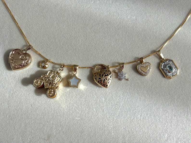 K&ouml;nnte beinhalten: Eine goldene Halskette mit verschiedenen Anh&auml;ngern, darunter ein herzf&ouml;rmiges Medaillon, eine Kutsche, ein Stern, ein herzf&ouml;rmiges Vorh&auml;ngeschloss, eine Blume, ein Herz mit goldenem Zentrum und ein rechteckiger Anh&auml;nger mit blauem Stein.