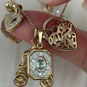 K&ouml;nnte beinhalten: Eine Sammlung goldfarbener Anh&auml;nger an filigranen Ketten. Die Anh&auml;nger umfassen ein herzf&ouml;rmiges Medaillon mit klaren Steinen, eine Kutsche mit einem ovalen blauen Edelstein und einen herzf&ouml;rmigen Anh&auml;nger mit floralen Ausschnitten.
