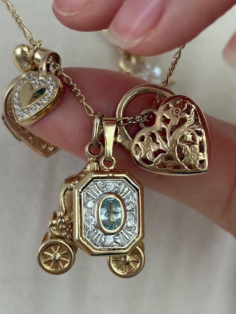 K&ouml;nnte beinhalten: Eine Sammlung goldfarbener Anh&auml;nger an filigranen Ketten. Die Anh&auml;nger umfassen ein herzf&ouml;rmiges Medaillon mit klaren Steinen, eine Kutsche mit einem ovalen blauen Edelstein und einen herzf&ouml;rmigen Anh&auml;nger mit floralen Ausschnitten.