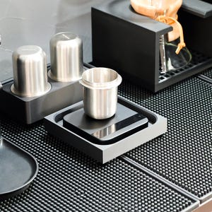 Könnte beinhalten: Eine moderne Kaffeestation mit einer schwarzen Mühle, Edelstahlbechern und einer Digitalwaage. Die Szene umfasst eine schwarze Abtropfschale und einen schwarzen Ständer für eine Pour-Over-Kaffeemaschine. Das Setup befindet sich auf einer strukturierten schwarzen Oberfläche.