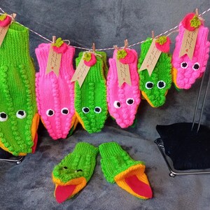 May include: A set of six knitted finger puppets, each with a different design. The puppets are green and pink with black eyes and a yellow mouth. The puppets are hanging on a wire rack.