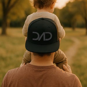 Könnte beinhalten: Schwarze Baseballkappe mit dem Wort "DAD" in grauen Buchstaben. Die Kappe wird von einer Person mit einem Kind auf den Schultern getragen. Der Hintergrund ist eine verschwommene Szene im Freien mit Bäumen und Gras.