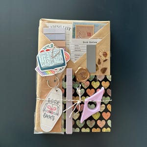 Puede incluir: Un paquete de papel marr&oacute;n envuelto con cuerda y decorado con varias pegatinas y ef&iacute;meras de papel. Las pegatinas presentan ilustraciones y texto con tem&aacute;tica de libros, incluyendo "The Book Was Better" y "Book Lover". El paquete tambi&eacute;n incluye un peque&ntilde;o marcap&aacute;ginas morado y un sello de cera dorado.