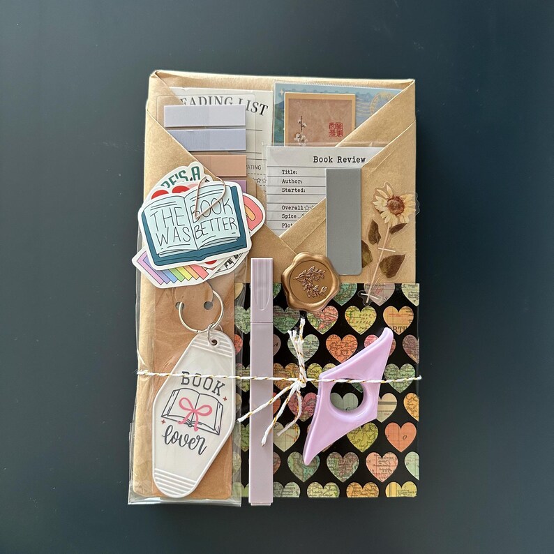 Puede incluir: Un paquete de papel marr&oacute;n envuelto con cuerda y decorado con varias pegatinas y ef&iacute;meras de papel. Las pegatinas presentan ilustraciones y texto con tem&aacute;tica de libros, incluyendo "The Book Was Better" y "Book Lover". El paquete tambi&eacute;n incluye un peque&ntilde;o marcap&aacute;ginas morado y un sello de cera dorado.