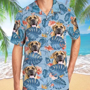 May include: A light blue short-sleeve button-down shirt featuring a repeating pattern of tan dog faces with open mouths and tongues out, interspersed with large dark blue monstera leaves and orange hibiscus flowers across the entire garment.