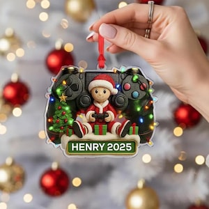 May include: A Christmas ornament shaped like a video game controller, featuring a Santa Claus figure holding a game controller. The ornament is decorated with a Christmas tree, presents, and the text "HENRY 2025".
