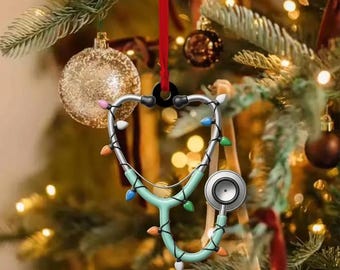 May include: A stethoscope Christmas ornament with a teal body and silver tubing, adorned with colorful lights. The ornament hangs from a red ribbon against a backdrop of a decorated Christmas tree with gold and brown ornaments.