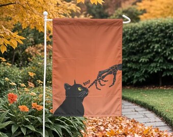 May include: Halloween garden flag with a black cat and skeleton hand design. The cat has yellow eyes and the word "BOOP" is next to its nose. The flag is orange and is hanging on a white pole.