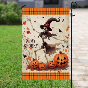 May include: Halloween garden flag featuring a skeleton wearing a witch hat and cape, with pumpkins and bats. The flag has the text "STAY SPOOKY" and a plaid border. The design is in orange, black, and beige.