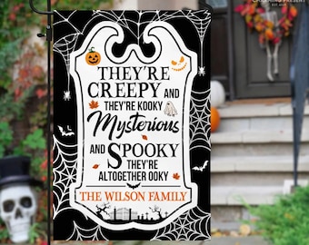 May include: A black and white Halloween garden flag with the text "They're Creepy and Kooky, Mysterious and Spooky". The flag features a pumpkin, ghost, and spiderweb design, with "The Wilson Family" at the bottom.