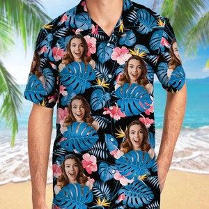 May include: A short-sleeve button-down shirt featuring a black background with a repeating pattern of blue monstera leaves, pink hibiscus flowers, and yellow bird of paradise flowers, interspersed with a repeating portrait of a smiling person.