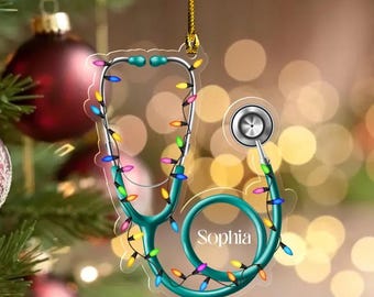 May include: A teal stethoscope Christmas ornament with colorful lights strung around it. The name "Sophia" is printed on the stethoscope. The ornament is hanging on a Christmas tree with red and gold ornaments.