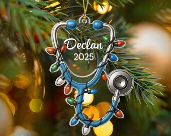May include: A Christmas tree ornament shaped like a stethoscope, with the name "Declan" and the year "2025" written on it. The stethoscope is blue and silver, adorned with colorful Christmas lights. The ornament hangs from a gold string.