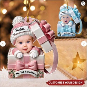 May include: Christmas ornament featuring a baby's face peeking out of a gift box. The box is white and pink with a large bow and the words "My 1st Christmas." The baby wears a pink hat with the name "Sophia" on it.