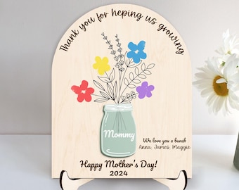 May include: Wooden sign with a hand-painted floral design and the text "Thank you for helping us growing". The sign also features a blue mason jar with the word "Mommy" on it and the text "We love you a bunch Anna, James, Maggie Happy Mother's Day! 2024".