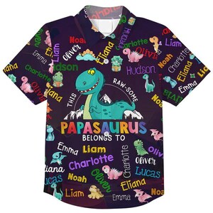 May include: A short-sleeved button-down shirt with a dark purple background, featuring a dinosaur theme. The shirt has a large green dinosaur graphic, the text "PAPASAURUS BELONGS TO", and various names in colorful text.