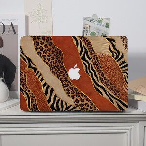 Könnte beinhalten: Eine Laptop-Hartschalenhülle mit einem gemusterten Design aus wellenförmigen vertikalen Abschnitten mit Leopardenmuster, Zebrastreifen und einfarbigen rostfarbenen Texturen, getrennt durch dünne goldene Linien, mit einem zentralen Ausschnitt für das Apple-Logo.