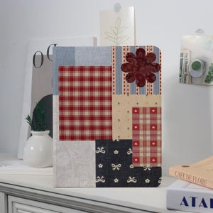 May include: A decorative notebook with a patchwork design. The cover features various fabric patterns, including red plaid, blue stripes, and floral prints. A large, burgundy flower is appliquéd on the top right corner. The notebook is rectangular and appears to be on a white surface.