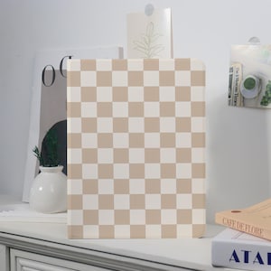 May include: A beige and white checkered pattern notebook or journal stands upright on a white surface. The cover features a grid of alternating squares. Other items on the surface include a vase with greenery, and books.