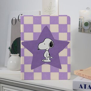 May include: A purple and white checkered notebook featuring a large purple star with a Snoopy character illustration. The notebook is standing upright on a white surface, with other books and decorative items in the background.