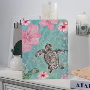 May include: A notebook with a watercolor design featuring a sea turtle swimming among pink hibiscus flowers and bubbles against a teal background. The notebook has a white border and is displayed on a white surface.