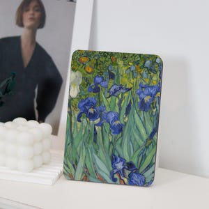 May include: A close-up of a painting of blue irises with green leaves and yellow flowers in the background. The painting is framed and displayed on a white surface.