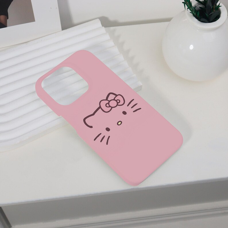 K&ouml;nnte beinhalten: Eine rosa Handyh&uuml;lle mit einem braunen Cartoon-Katzen-Gesicht-Design. Die H&uuml;lle hat einen Kameraausschnitt und liegt auf einer wei&szlig;en Oberfl&auml;che. Das Katzengesicht hat eine Schleife, Schnurrhaare und eine Nase.