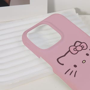 K&ouml;nnte beinhalten: Eine hellrosa Handyh&uuml;lle mit einem Cartoon-Katzen-Gesicht-Design. Die H&uuml;lle hat einen Ausschnitt f&uuml;r die Kamera und eine glatte Oberfl&auml;che. Das Katzendesign umfasst eine Schleife, Schnurrhaare und kleine Punkte.