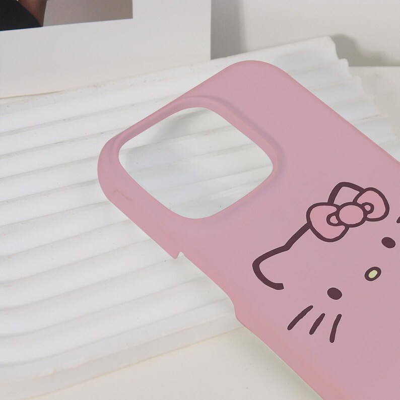 K&ouml;nnte beinhalten: Eine hellrosa Handyh&uuml;lle mit einem Cartoon-Katzen-Gesicht-Design. Die H&uuml;lle hat einen Ausschnitt f&uuml;r die Kamera und eine glatte Oberfl&auml;che. Das Katzendesign umfasst eine Schleife, Schnurrhaare und kleine Punkte.