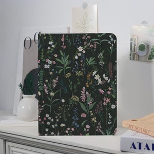 May include: A rectangular notebook with a dark background and a floral pattern. The design features a variety of colourful wildflowers and green leaves. The notebook is placed on a white surface.