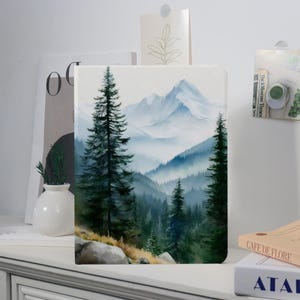 May include: A landscape painting of a mountain range with evergreen trees in the foreground. The artwork features a watercolor style with shades of blue, green, and white. The painting is displayed on a white surface.