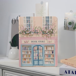 May include: A decorative notebook featuring a detailed illustration of a quaint book store. The facade is painted in soft pink and blue hues, with floral accents and the text "BOOK STORE" above the entrance. The notebook is standing on a white surface.