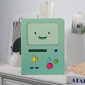 May include: A mint green tablet case featuring a cartoon face with a smiling mouth and black eyes. The case has a rectangular design with a screen and control buttons in yellow, blue, green, and pink. The case is on a white surface.