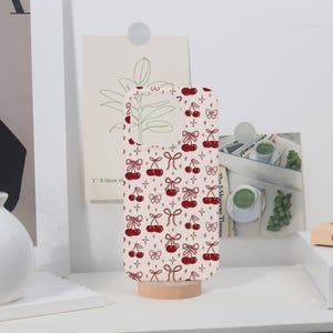 May include: A phone case with a cream background and a repeating pattern of red cherries and bows. The case is propped up on a small wooden stand, showcasing its design. The case has a cutout for the camera.