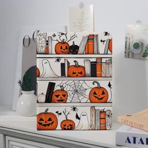 May include: A decorative Halloween-themed bookshelf illustration. The design features orange jack-o'-lanterns, books, spiders, and ghosts on shelves. The color palette includes orange, black, and white, creating a spooky yet charming aesthetic.