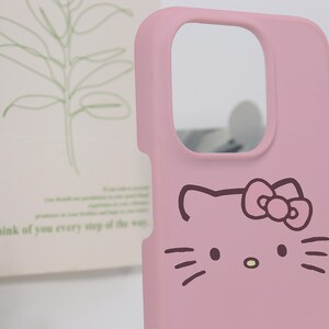 K&ouml;nnte beinhalten: Eine rosa Handyh&uuml;lle mit einem Cartoon-Katzen-Gesicht-Design. Die H&uuml;lle hat einen Kameraausschnitt und eine glatte Oberfl&auml;che. Das Katzengesicht hat Schnurrhaare, eine Nase und eine Schleife.