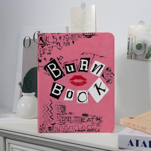 Könnte beinhalten: Ein pinkfarbenes Notizbuch mit den Worten "BURN BOOK" im Collage-Stil, mit einer roten Lippen-Grafik. Das Cover ist mit schwarzen Kritzeleien und Sätzen wie "HAIRY" und "EAT ME" verziert.