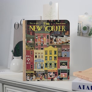 May include: A hardcover notebook featuring a vintage cover of The New Yorker magazine from March 18, 1944. The cover art depicts a colorful street scene with shops and people. The notebook is displayed on a white surface.